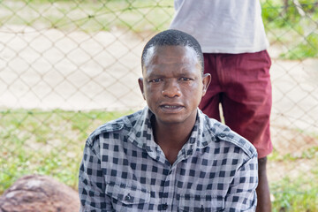 portrait of african man villager siting in the yard in front of a mesh fence outdoors , village life