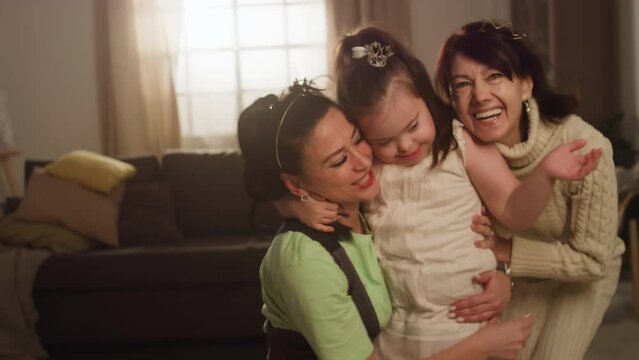 Portrait of happy three-generation family of mother, grandmother and down syndrome child, all smiling while posing in living room and looking at camera