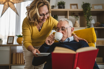 Senior man read book while his wife bring him cup of coffee at home