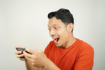 Crazy, funny and excited face of Asian man in orange t-shirt addicted to play mobile game