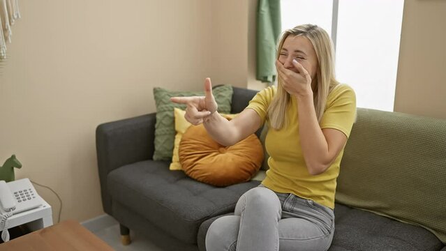 Cheeky young woman points to you in childish jest! gorgeous blonde laughing, sporting a t-shirt, hand at mouth in a playful expression of fun, all in the comfort of her cozy living room
