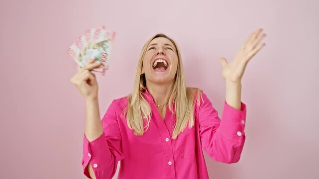 Joyful young blonde woman triumphantly holding new zealand dollars, ecstatically celebrating her win with a beaming, winner expression over an isolated pink background
