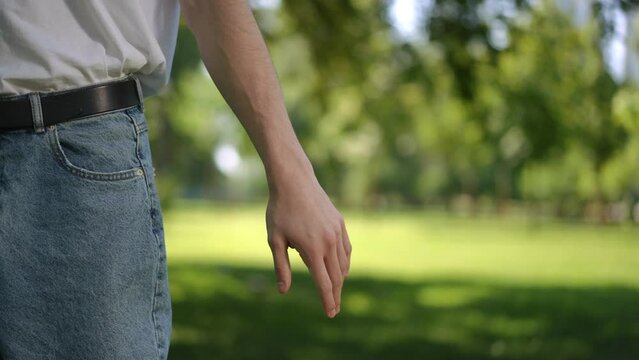 Slow motion. Close-up. An unrecognizable man, wearing a white T-shirt and jeans, clutches an empty plastic bottle in his hand and throws it on the grass as he walks through a city park on a sunny