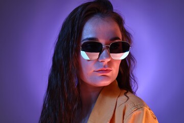 Fototapeta premium Portrait of beautiful young woman with stylish sunglasses on color background with neon lights