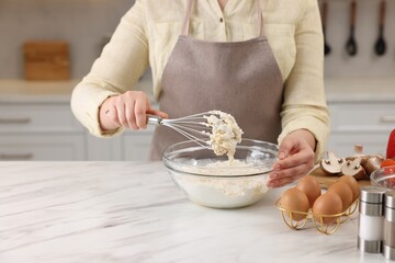 Woman making dough with whisk in bowl at light marble table, closeup