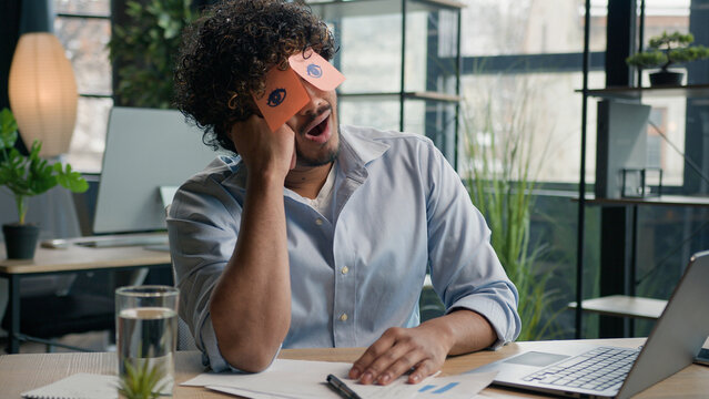 Funny lazy worker napping at office tired Arabian Indian business man sleeping covering eyes with sticky notes comical stickers on face employee businessman yawning pretend working cheating to sleep