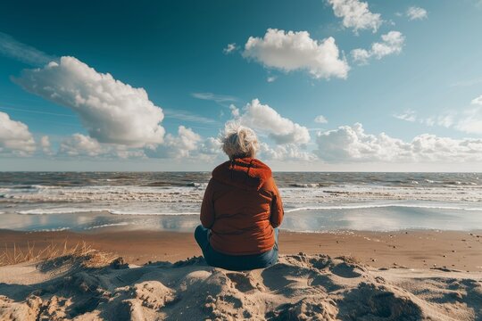 A senior woman wrapped in a warm jacket, sitting on the sand and looking out towards a cloudy beachscape