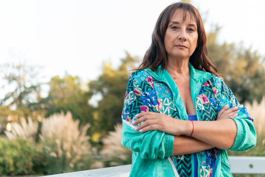 Beautiful senior woman 50 to 60 years old, wearing colorful clothes, 80s style, posing, arms crossed confidently, after doing exercises, in an outdoor park at sunset