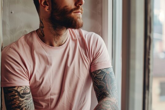 A tattooed man with a beard sits by the window in a pink t-shirt and looks out the window