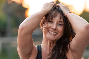 Beautiful adult woman 50 to 60 years old smiling with joy, raising her arms to celebrate, wearing sports clothing in a park at sunset.