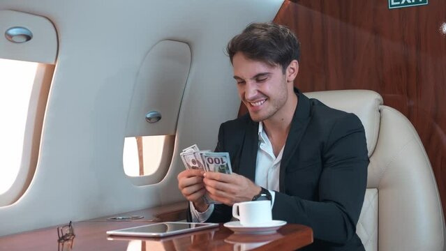 Young billionaire on board his private jet sits at table and counts cash. Billionaire laughs happily and throws bills in air, enjoying financial success. Billionaire wealth concept on private jet