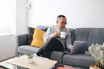 Senior Asian man sitting on sofa and using laptop while holding a cup of coffee in a modern living room