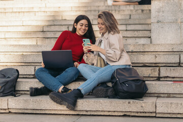 Two student girls sitting on the stairs of the college with the laptop and the mobile