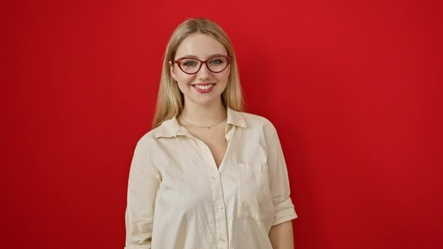 Blonde bombshell in specs, adorable young woman playfully sends love, blowing a kiss to the camera over isolated red backdrop