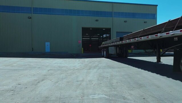 Low angle dolly follows semi truck with steel beams reversing into warehouse at midday