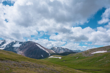 Beautiful view to green alpine valley among hills and big rocky ridge with snow under clouds in blue sky. Shadows of clouds on hilly terrain in changeable weather. Large wide mountain range far away.