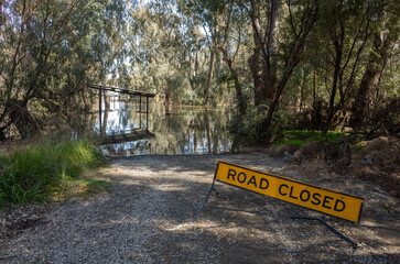 A 'ROAD CLOSED' sign is placed on a submerged footpath along the riverbank, indicating an...