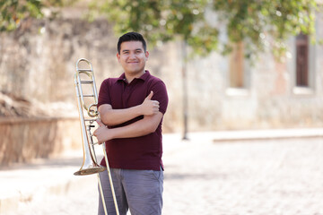 portrait of young mexican man smiling with thumbs up and holding trombone with blurred background