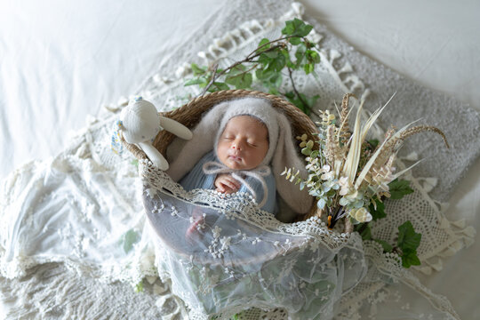 Newborn photo of a newborn baby who is 8 days old, half Taiwanese and half Australian, wrapped in a blue wrap, wearing a rabbit hat and sleeping in a basket. 生後8日の台湾人とオーストラリア人のハーフの新生児の赤ちゃんが青いおくるみを巻かれて