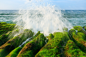 Tidal waves splash up radial water sprays when crashing on the shore of massive green rocky...