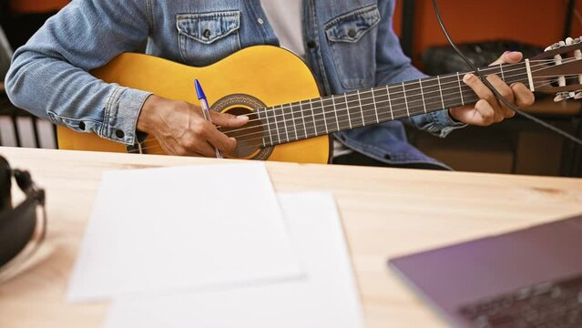 A young hispanic man writing music in a studio with a laptop and guitar.