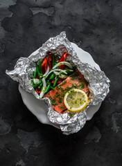 Lunch preparation - salmon baked with vegetables in foil on a dark background, top view