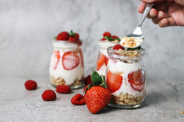 Hand holding spoon with parfait made from strawberries, yogurt and granola over grey background 