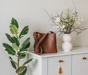 A brown leather tote bag, a vase with flowers on a white chest of drawers, a ficus in a basket in...