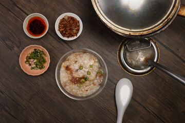 Asian thick rice porridge bubur chicken beef meat fish prawn rustic silver pot ladle serving in glass bowl fried shallots green onion chilli oil condiment