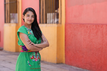 Fototapeta premium Portrait of smiling young Mexican girl dressed in typical Mexican clothing, looking at the camera with crossed arms with out-of-focus street background.