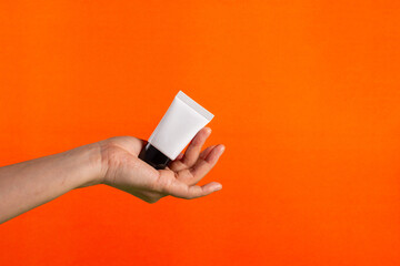 Plastic white tube for cream or lotion. Skin care or sunscreen cosmetic in hand on orange background.