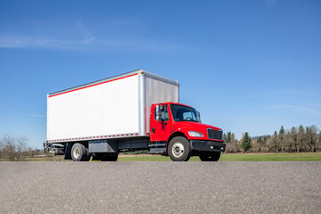 Bright red middle duty day cab rig semi truck transporting cargo in box trailer driving on the local road for delivery