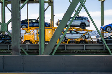 Bright yellow car hauler big rig semi-truck transporting cars on hydraulic semi trailer running on the truss metal bridge