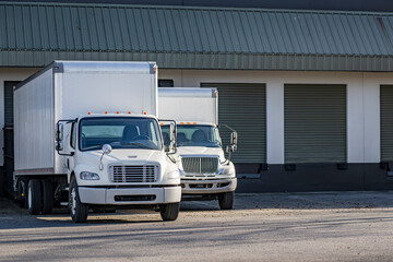 Day cab white middle duty rigs semi trucks with long box trailers standing in warehouse dock gates...