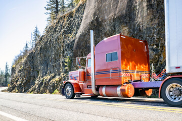 Extended cab orange big rig classic semi truck transporting cargo in dry van semi trailer driving on the mountain road with rocks and trees