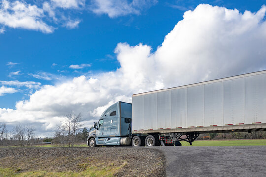 Light blue big rig industrial semi truck transporting cargo in dry van semi trailer driving on the local narrow road with cloudy sky on the background