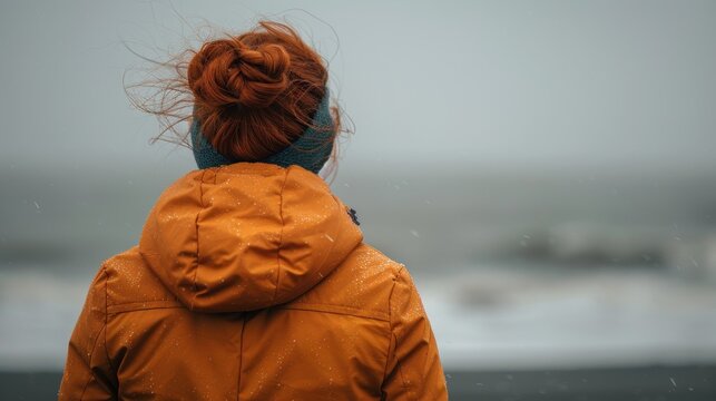 A woman in an orange jacket stands by the ocean, gazing into the distance