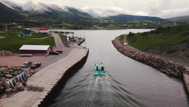 Drone shot following a small commercial lobster fishing boat coming in after a long day revealing a beautiful small village with mountains in the background on Cape Breton Island, Nova Scotia.