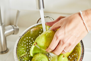 Woman in white shirt washing green pears under clear water in stainless steel kitchen sink