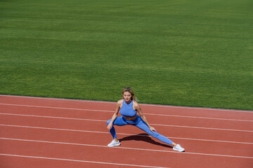 Fototapeta premium Female runner stretching before workout. Sports exercises and stretching in the stadium. Physical training girl. Fit girl do outward lunging. Physical training education. Sports lesson