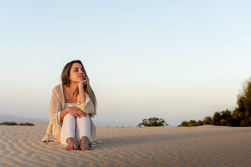 A woman sits peacefully on a sandy dune, her gaze thoughtfully directed towards the horizon as the...