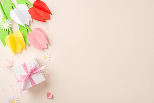 Creative Mother's Day concept, from top view of origami tulips, chamomiles, a DIY present box, ribbon-tied, alongside paper hearts and fine confetti on a pastel beige field