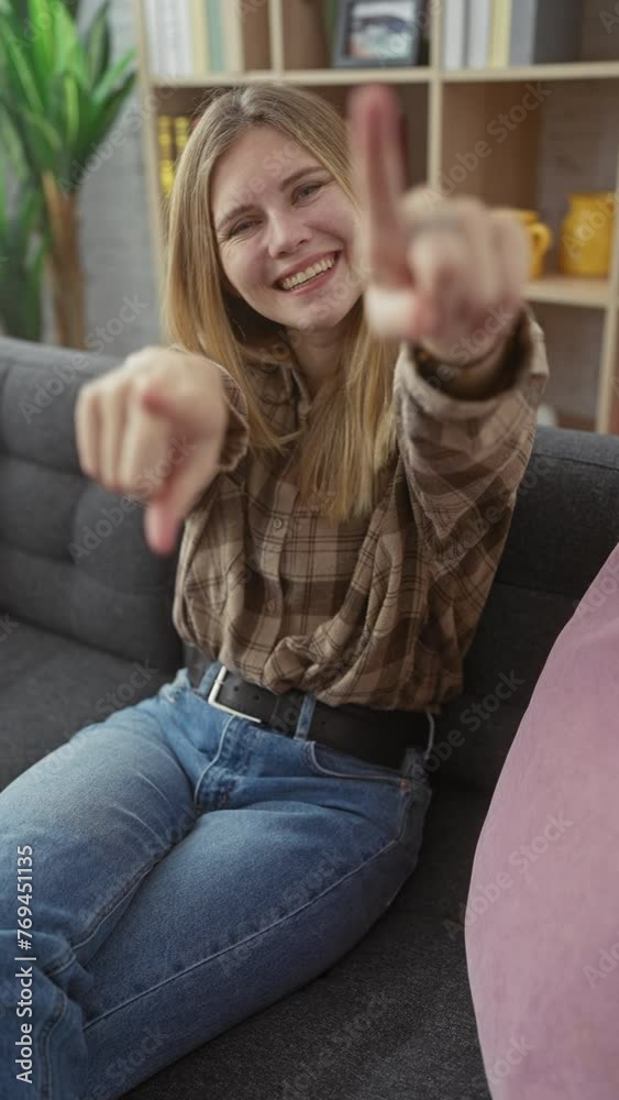 Sticker cheerful young blonde woman, casually sitting on her living room sofa, playfully pointing straight a