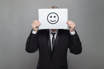 Anonymous businessman hiding his head behind happy smile emoticon on gray background - 769475105