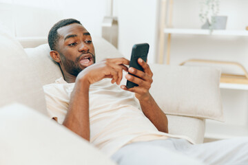 Happy African American man sitting on a black sofa, using his mobile phone He is relaxed and...