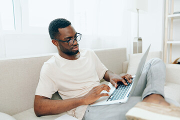 Smiling African American Man Typing on Laptop, Working Freelancer on Sofa in Modern Living Room A...