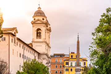 Naklejka premium The Trinita dei Monti church stands above the Roman skyline, bathed in the warm glow of a sunset, with an ancient obelisk adding contrast against the pastel-colored buildings.
