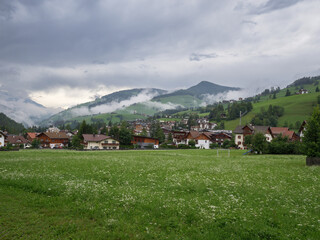 Green Meadows, Wooden Houses in the Village of San Vigilio di Marebbe and Italian Alps Mountains in the Background on a Cloudy Summer Day, Italy