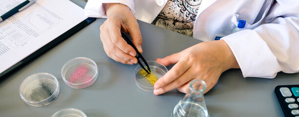 Banner of unrecognizable female scientist with tweezers examining golden glitter sample over petri dish on research laboratory. Woman chemist technician studying dangers of microplastics composition