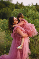 Naklejka premium Cheerful mother and her daughter in tulle pink dresses posing outdoor at summer. Beautiful mom holding her little girl in her arms on nature background. Photoshooting